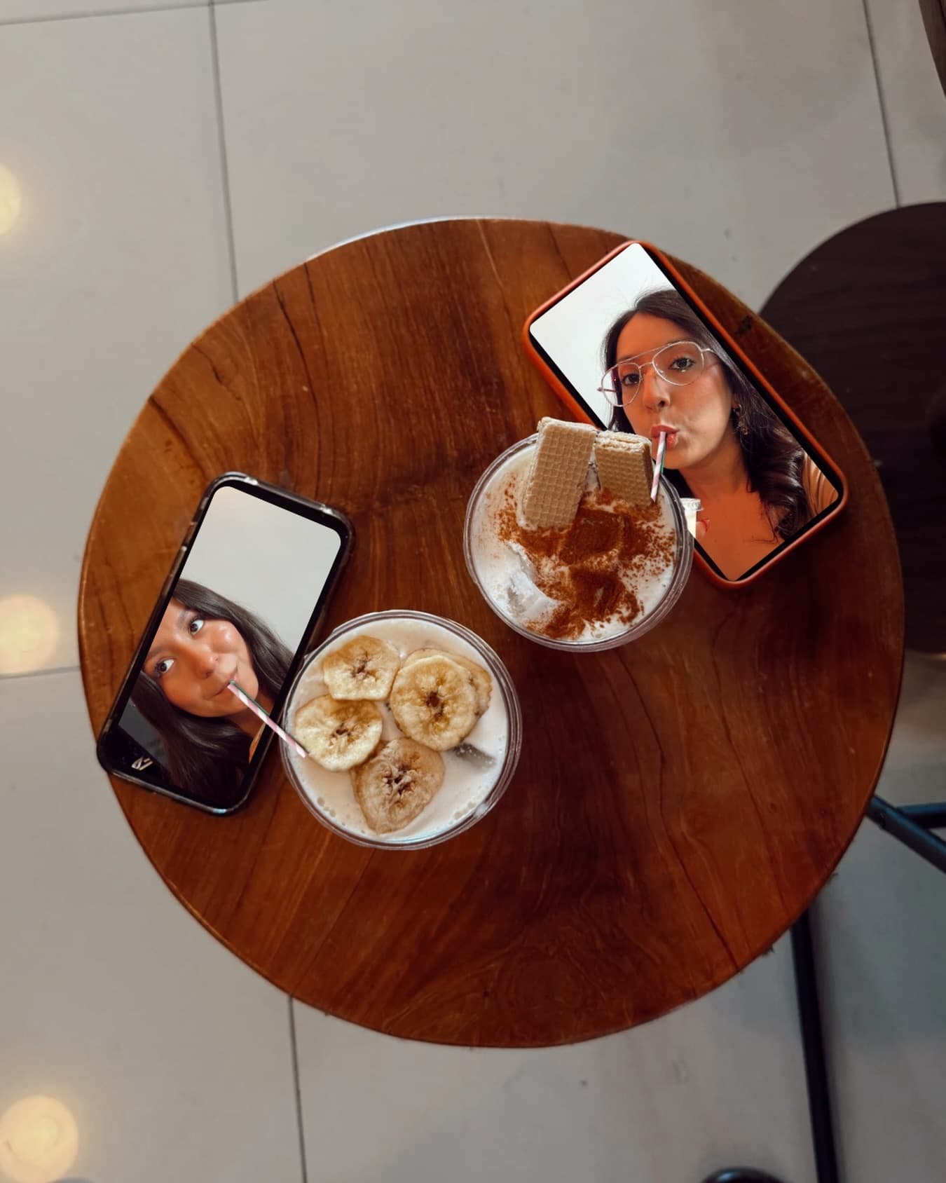 Overhead view of an iced banana latte and a cinnamon-wafer latte on a round wood table, flanked by two phones showing friends sipping.