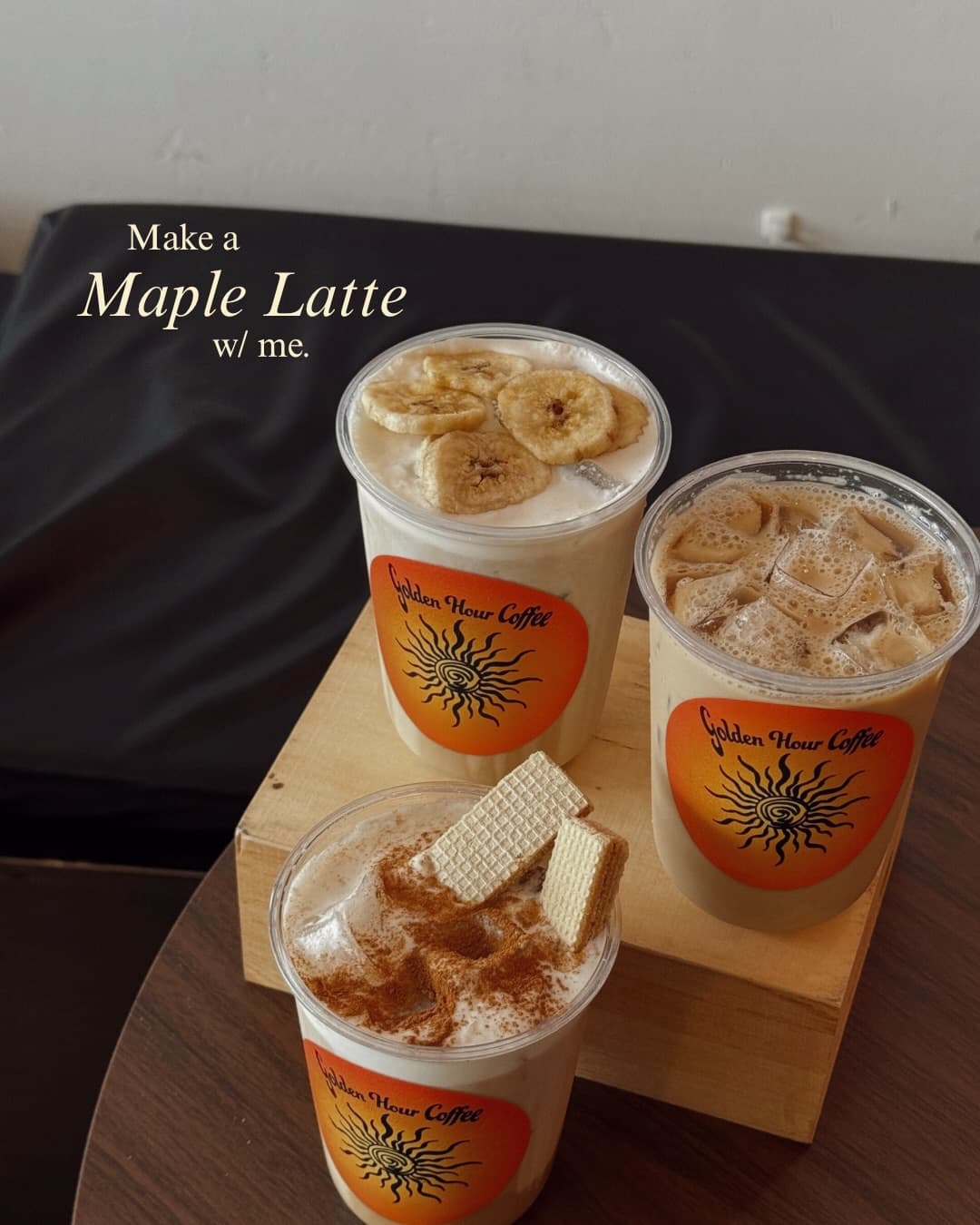 Three Golden Hour drinks staged on a wooden riser against a dark wall: a banana-chip latte, an iced coffee, and a cinnamon-dusted latte with wafer cookies.