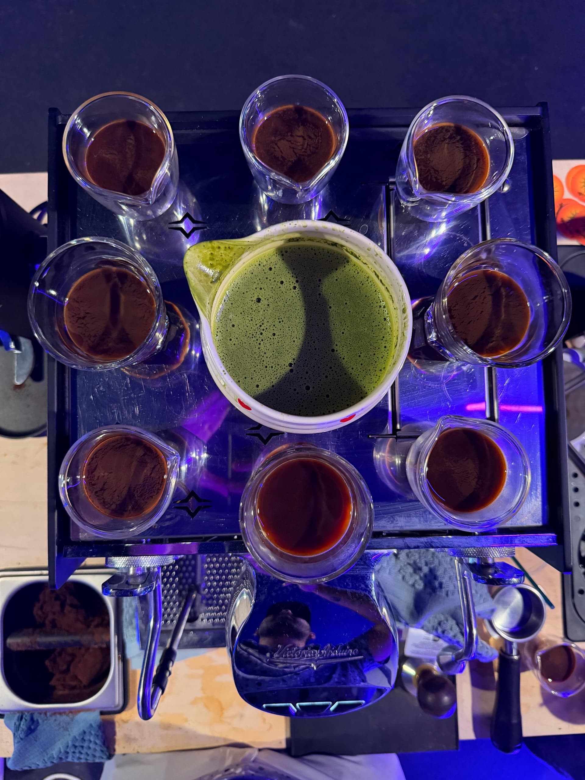 A top-down view of fresh espresso shots surrounding a pitcher of whisked matcha on the coffee bar.