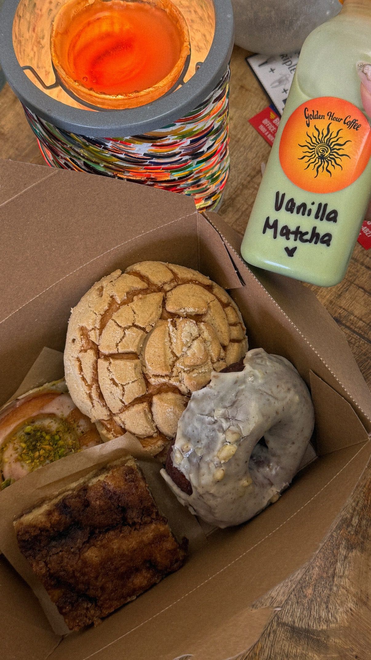 A green Vanilla Matcha bottle with the orange sun label beside a kraft box of pastries, lit by warm candlelight.